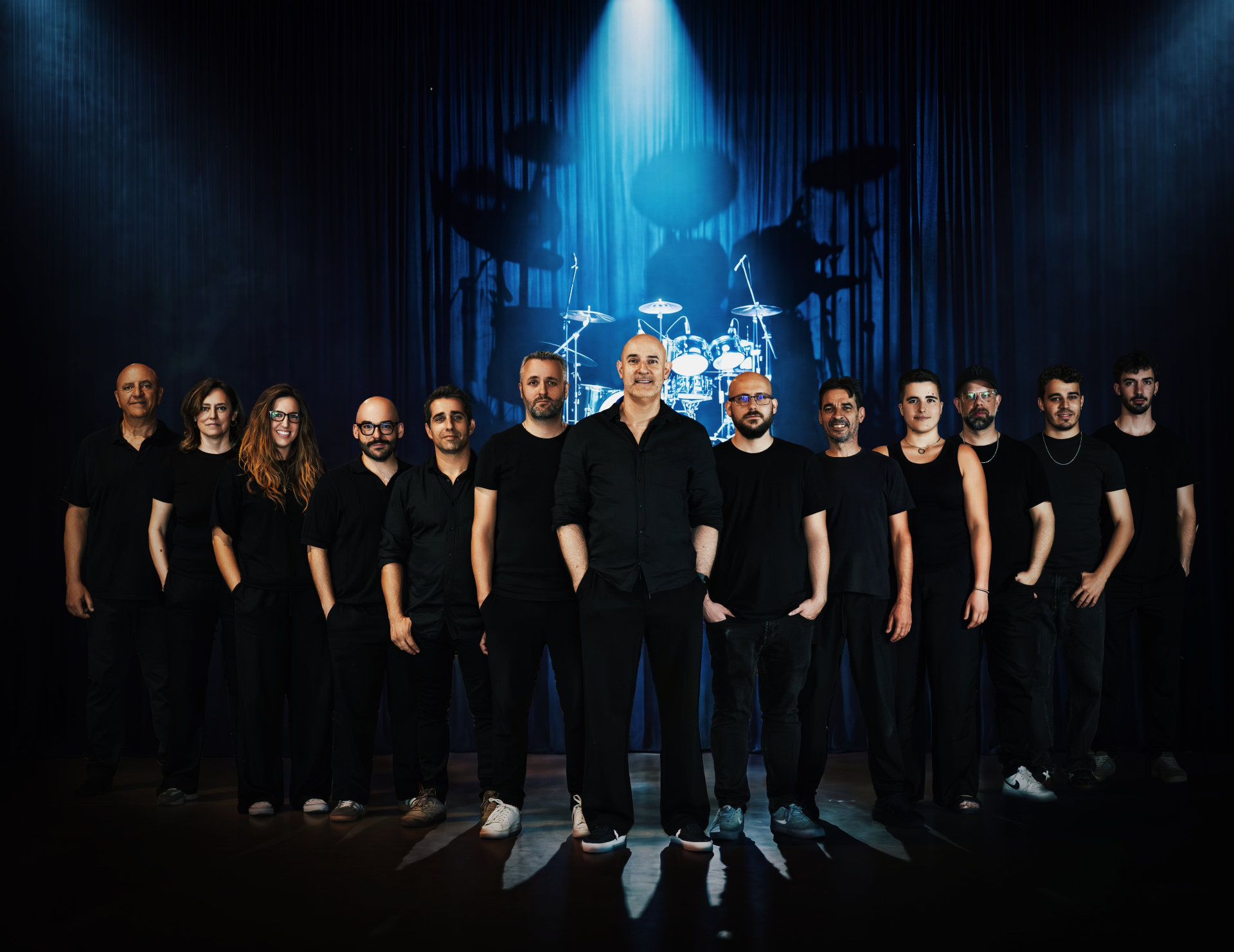 The 13 musicians of IN THE AIR TO BAND on stage under a blue spotlight with drums in the background